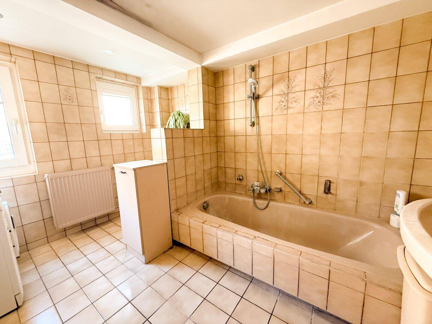Badezimmer mit beigen Fliesen, Badewanne und Fenster für natürliche Belüftung.