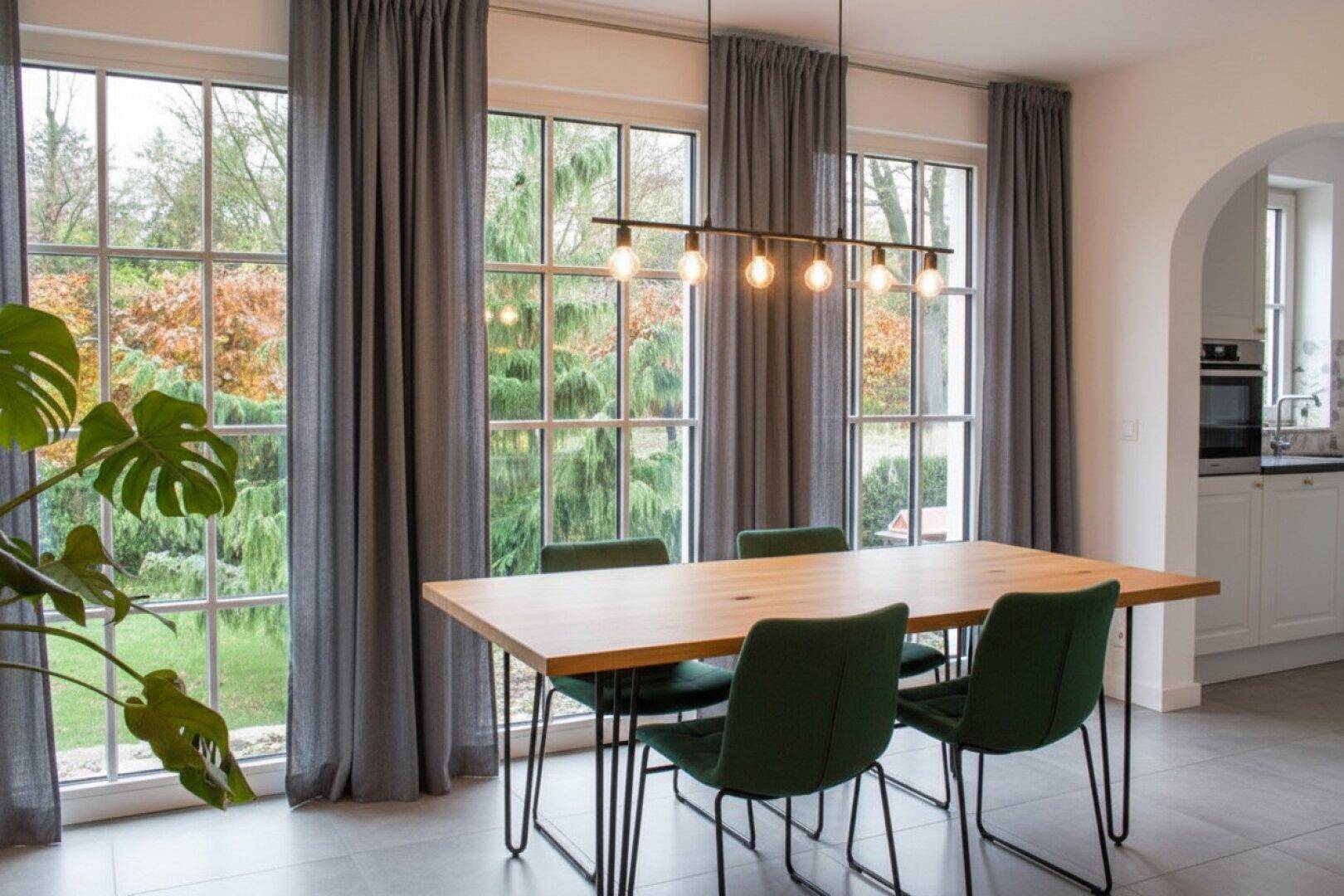 Moderner Essbereich mit Holztisch, grünen Stühlen und großen Fenstern mit Blick in den Garten.