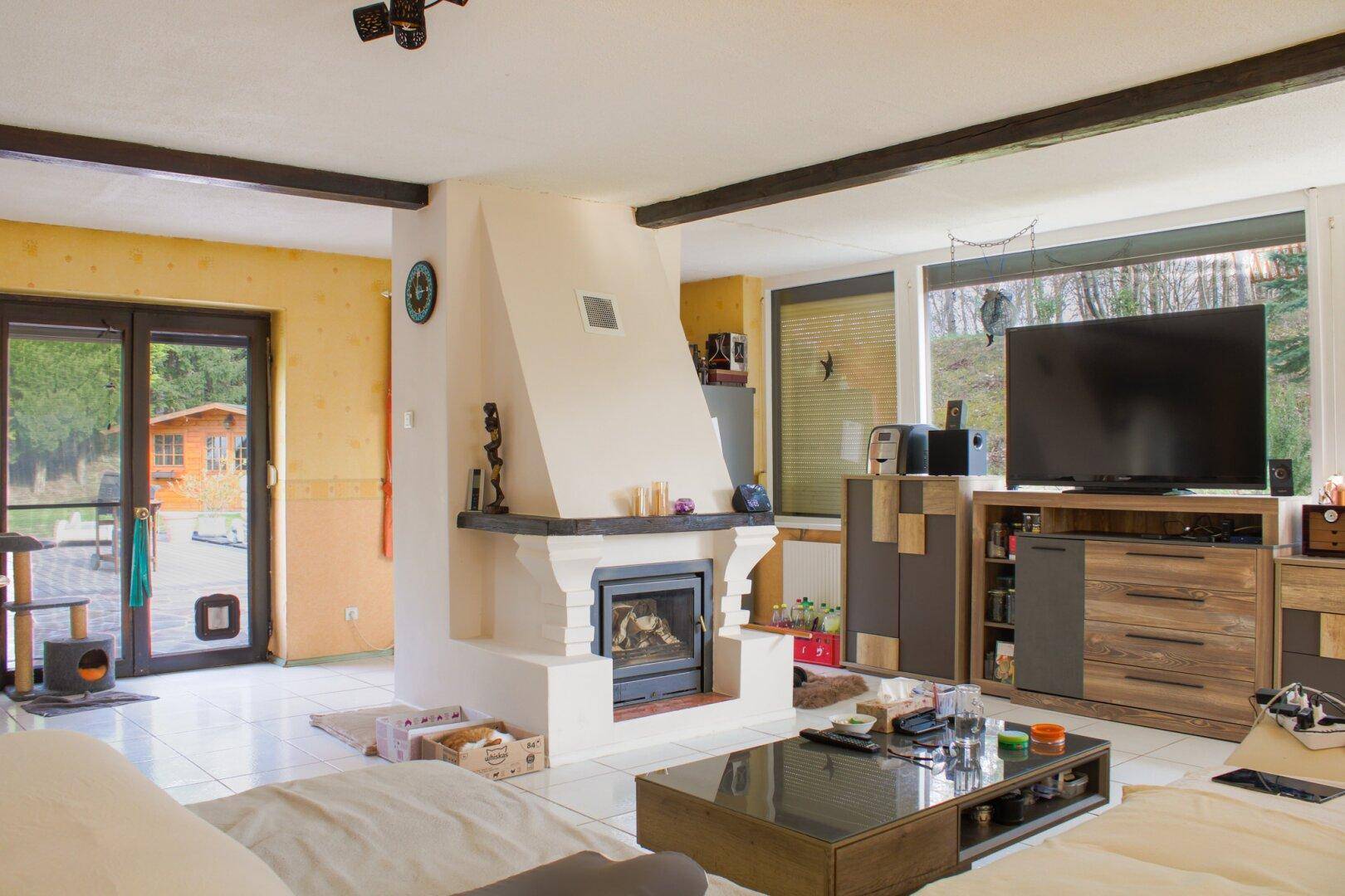Helles Wohnzimmer mit Kamin, Fliesenboden und Blick ins Grüne durch große Fenster.