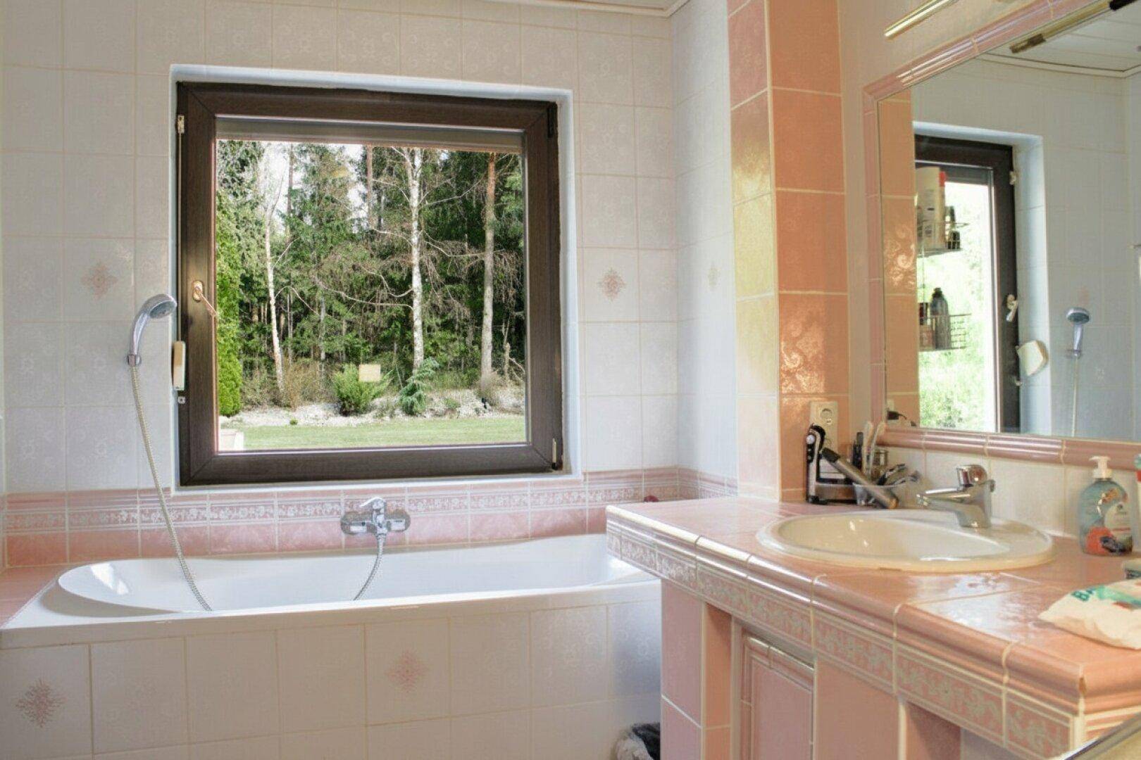 Badezimmer mit Badewanne, rosa Fliesen und großem Fenster mit Blick ins Grüne.