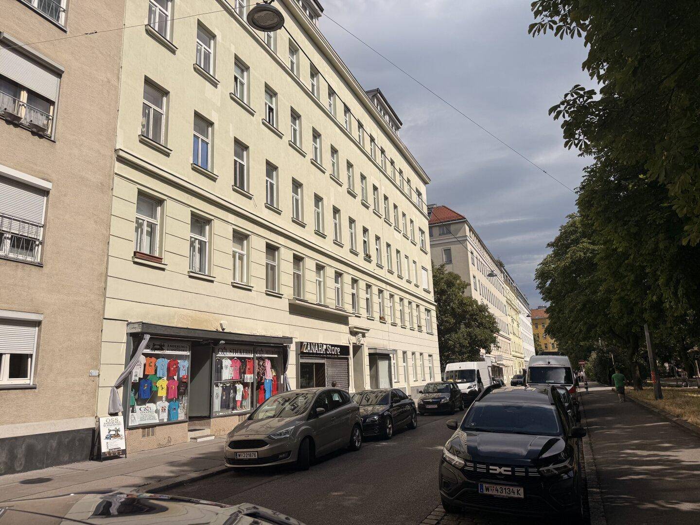 Mehrstöckiges Wohn- und Geschäftshaus mit Ladenlokal im Erdgeschoss und parkenden Autos auf der Straße.
