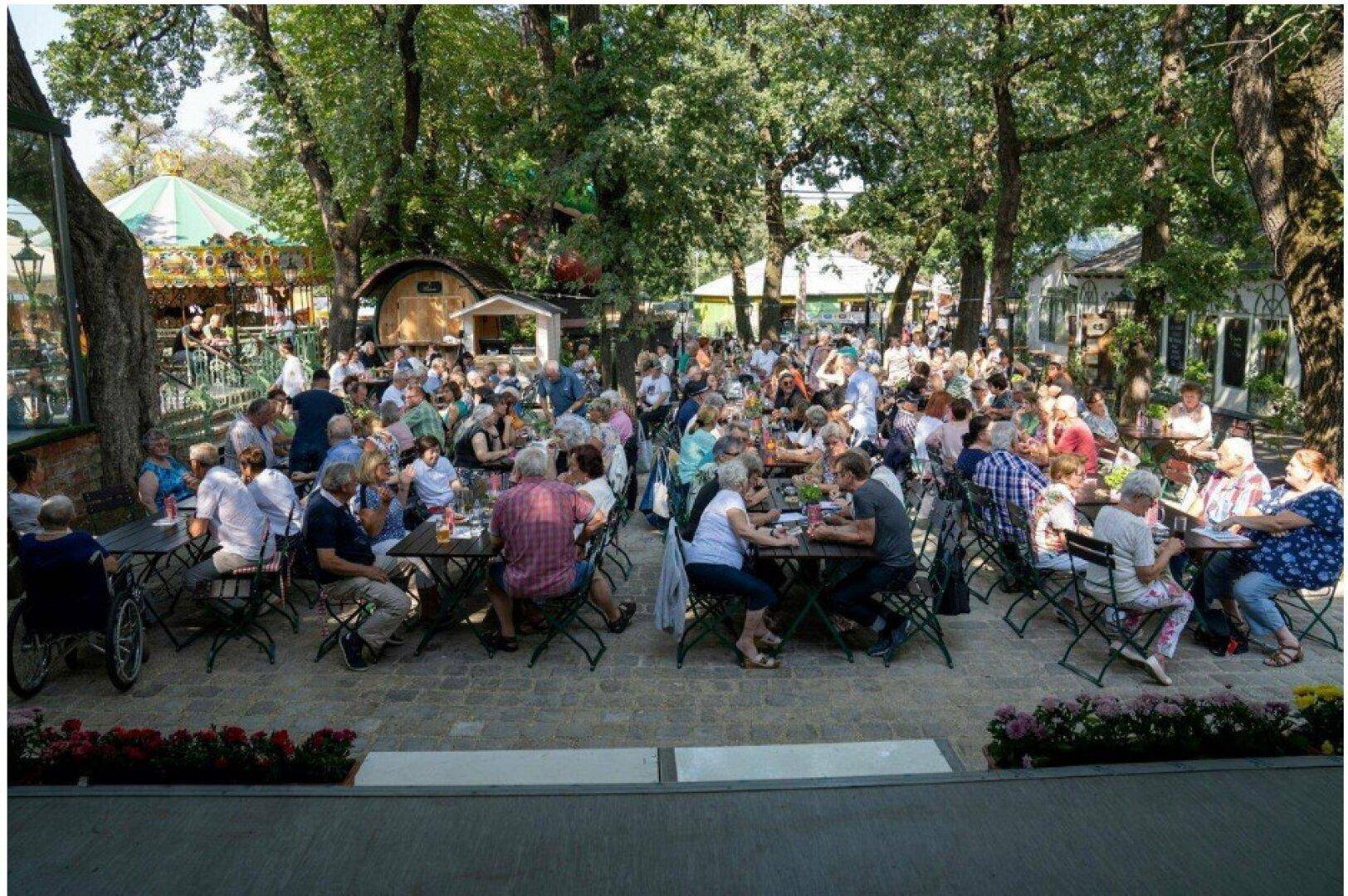 Belebtes Gartenrestaurant mit zahlreichen Gästen unter hohen Bäumen, ideal für gesellige Stunden im Freien.