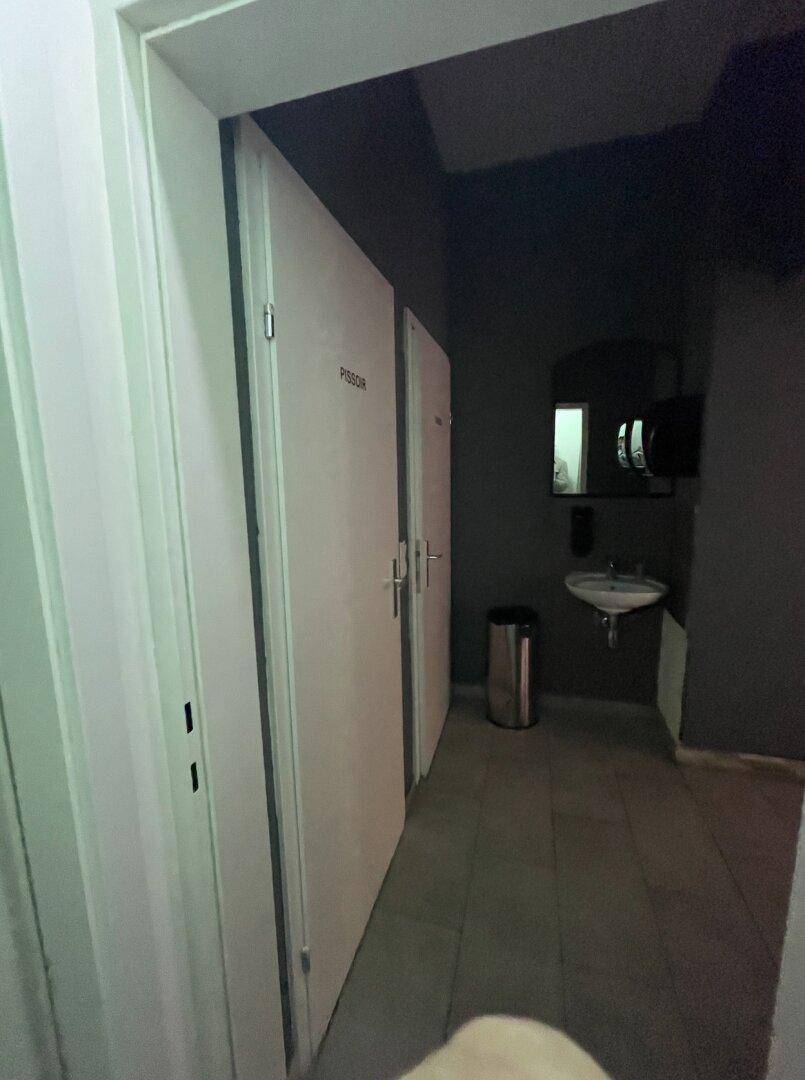 Schmaler Flur zu den Toiletten mit Waschbecken und Spiegel, in dunklen Farbtönen gehalten.