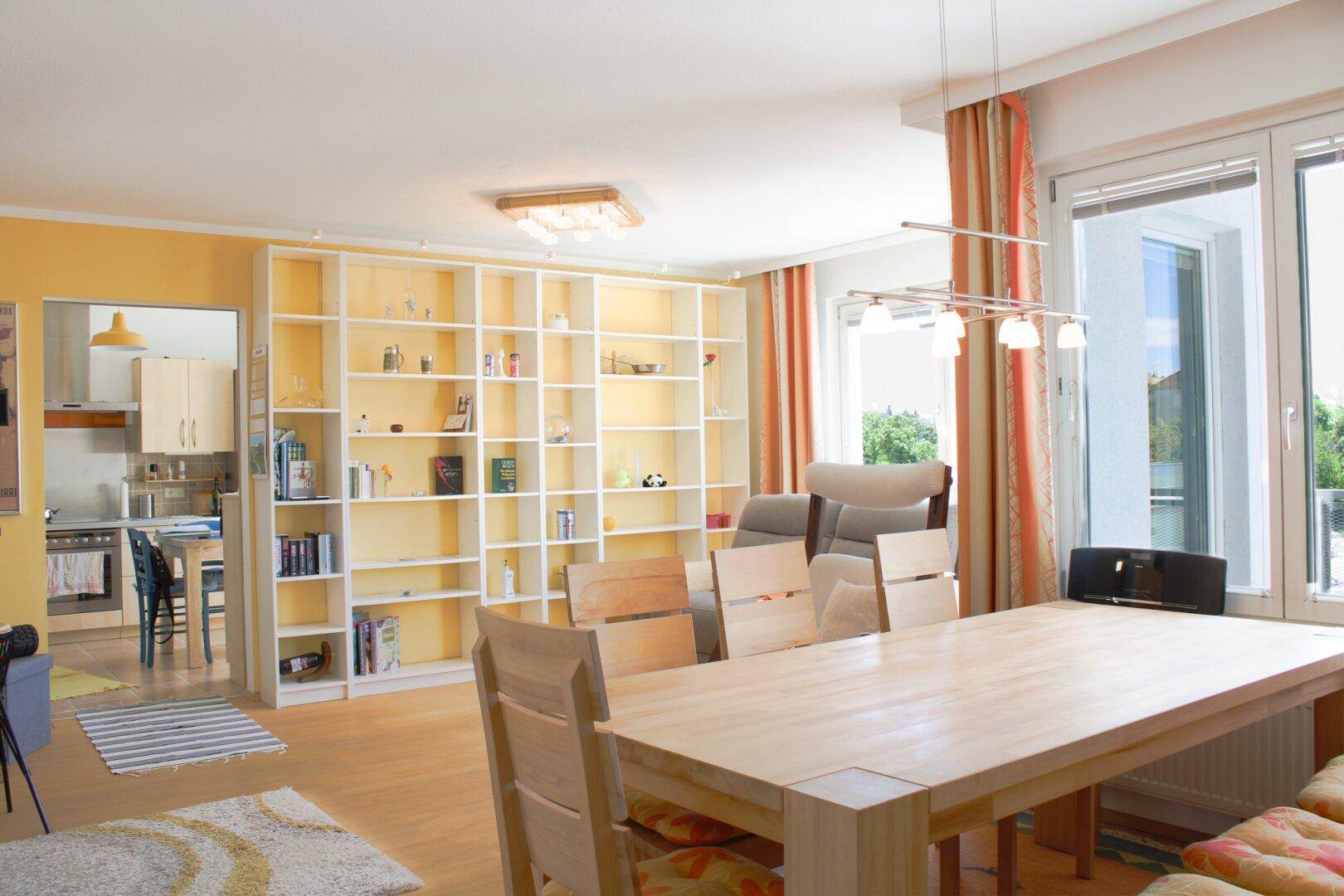 Geräumiger Wohn-Essbereich mit hellem Holztisch, großem Bücherregal und Zugang zum Balkon.