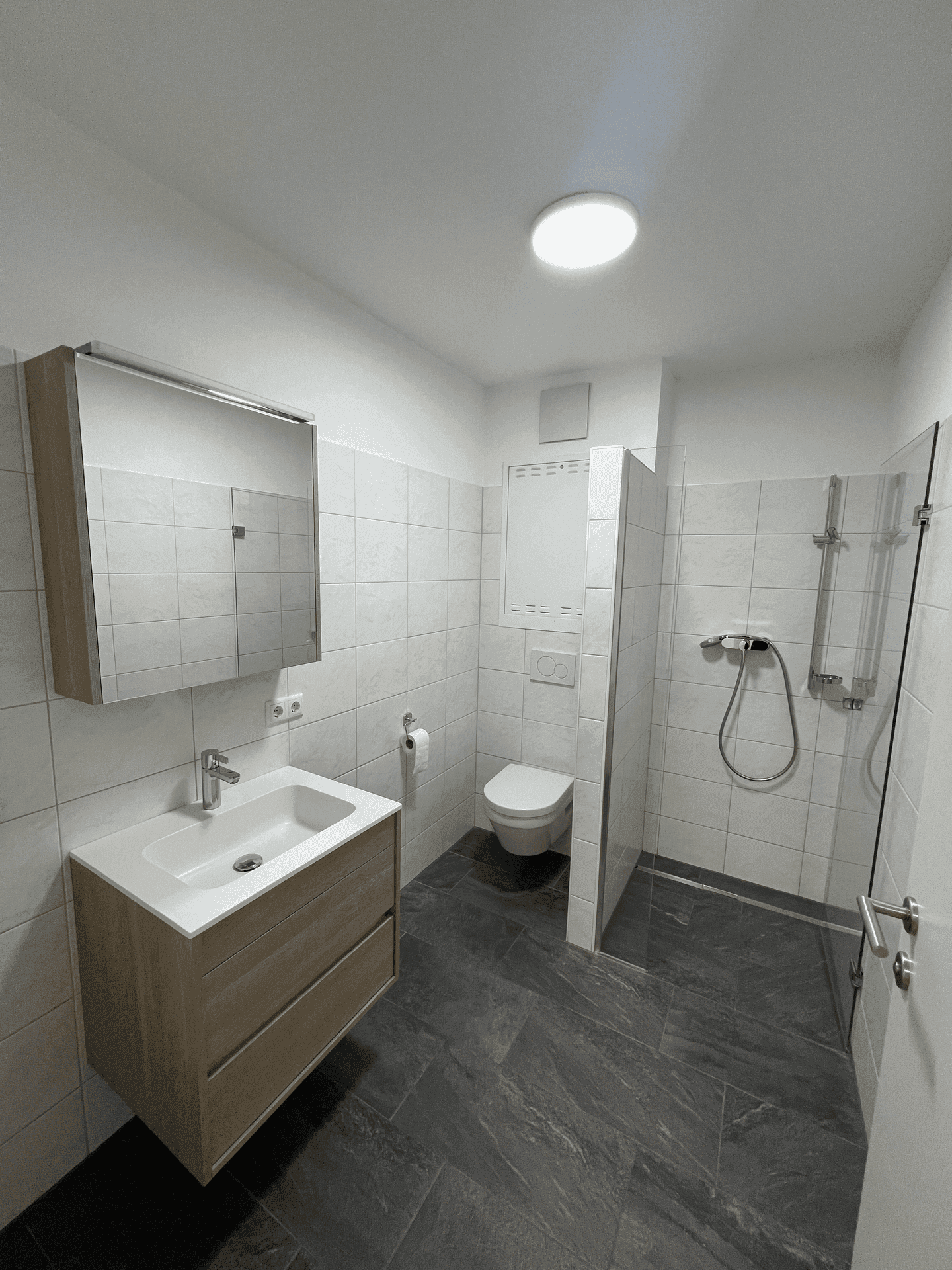 Modernes Badezimmer mit ebenerdiger Dusche, Waschbecken und dunklen Bodenfliesen.