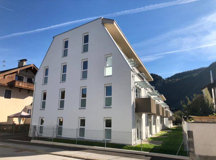 Modernes Mehrfamilienhaus mit weißer Fassade und Balkonen in einer bergigen Landschaft.
