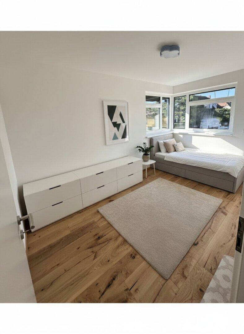 Helles Schlafzimmer mit Holzboden, großem Fenster und minimalistischer Einrichtung.