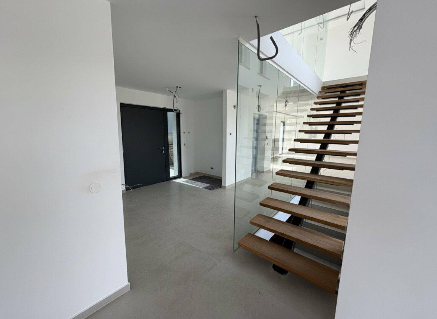 Eingangsbereich im Bau mit moderner Holztreppe, Glasgeländer und großzügigem Raumgefühl.
