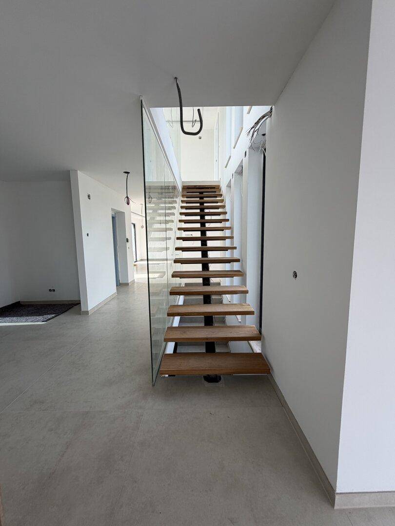 Moderne Holztreppe mit Glasgeländer in einem hellen, noch unfertigen Innenraum.