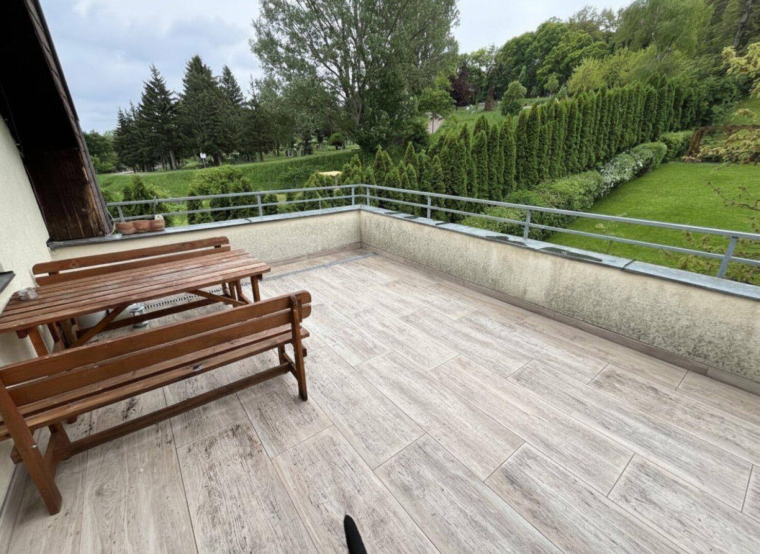 Geräumige Terrasse mit Holztisch und Bänken, umgeben von grüner Landschaft und Bäumen.