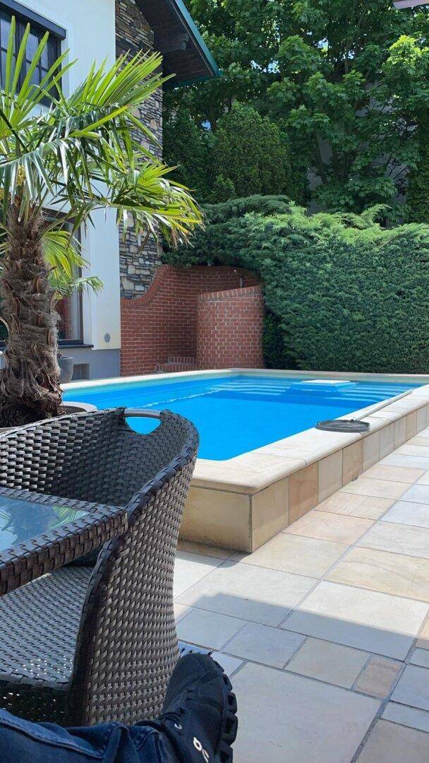 Blick auf den Swimmingpool und die Terrasse mit Korbmöbeln, umgeben von Palmen und üppigem Grün.