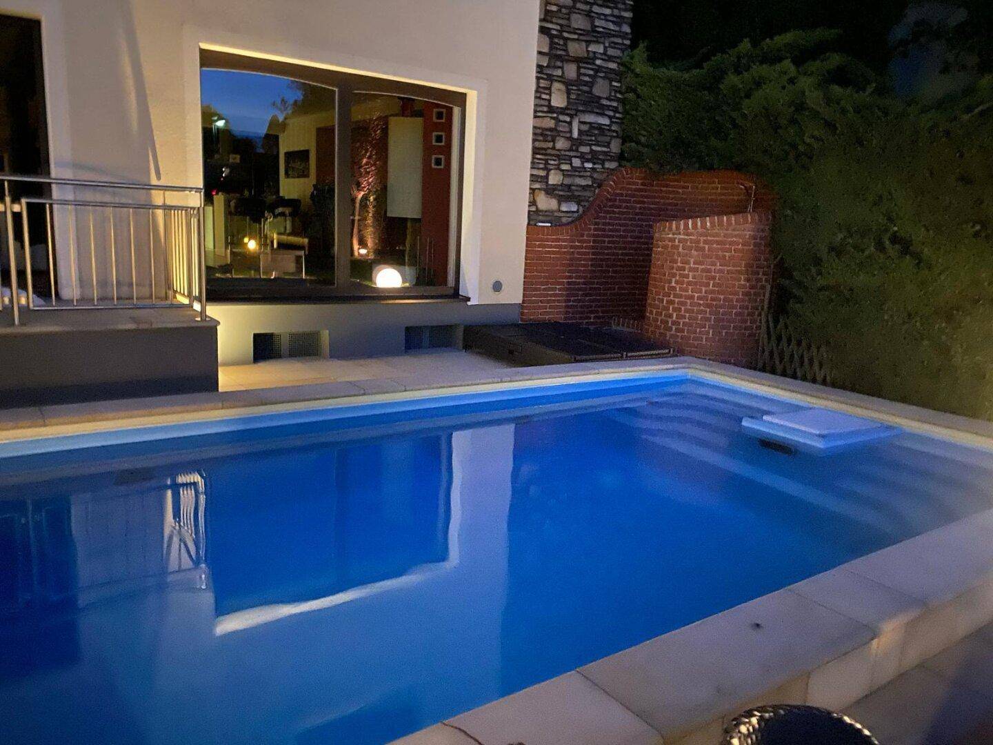Beleuchteter Swimmingpool im Außenbereich bei Nacht, mit reflektierenden Lichtern auf der Wasseroberfläche.