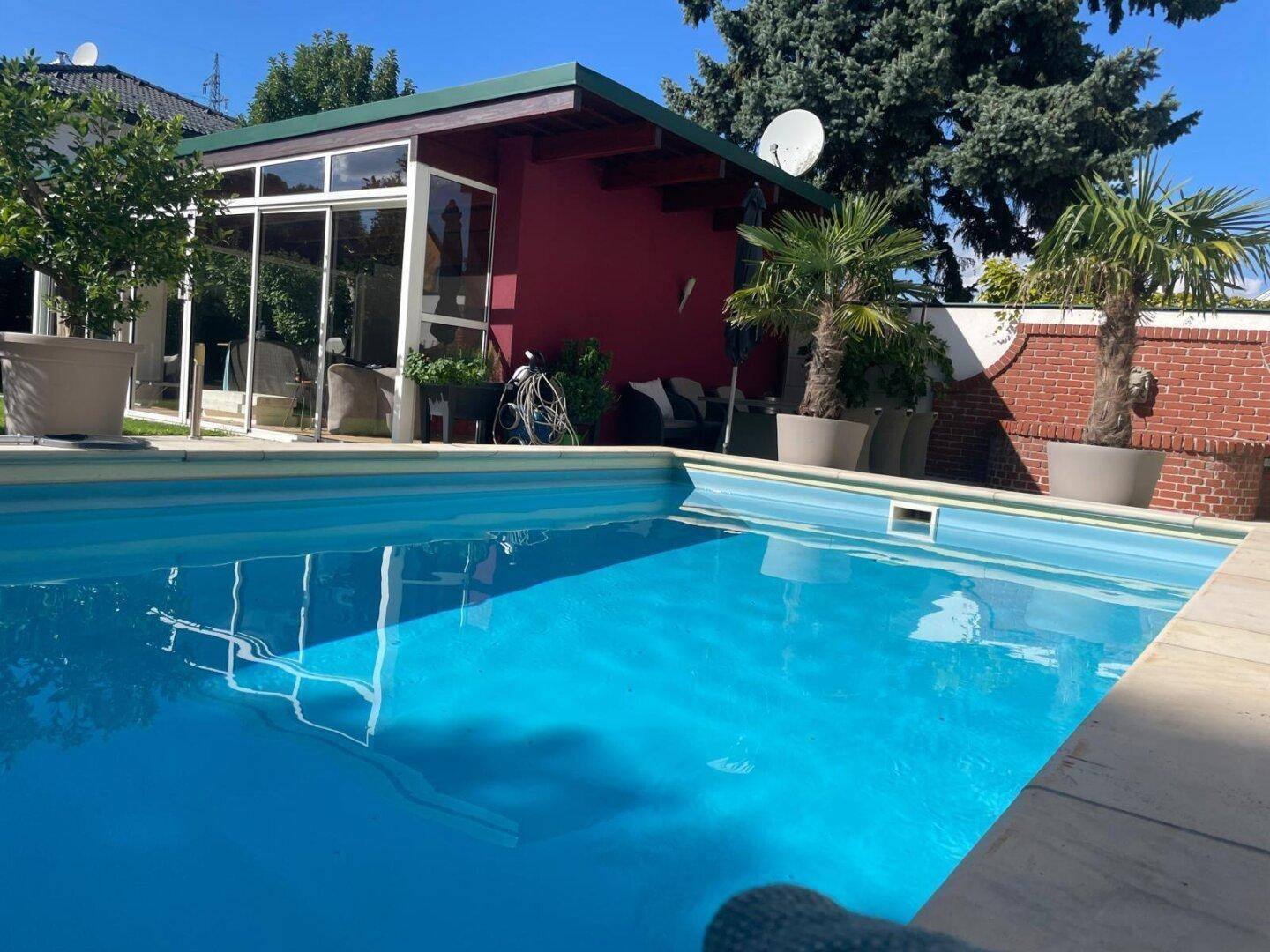Außenbereich mit großem, blauem Swimmingpool und einem modernen Poolhaus unter strahlend blauem Himmel.
