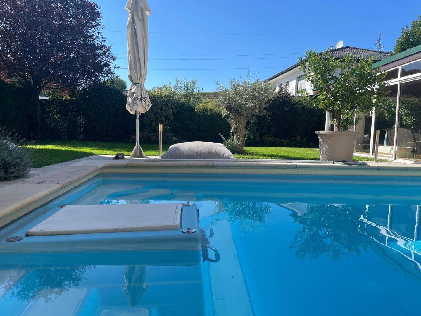 Großer Swimmingpool im Gartenbereich mit Rasenfläche, Bäumen und Sonnenschirm an einem sonnigen Tag.