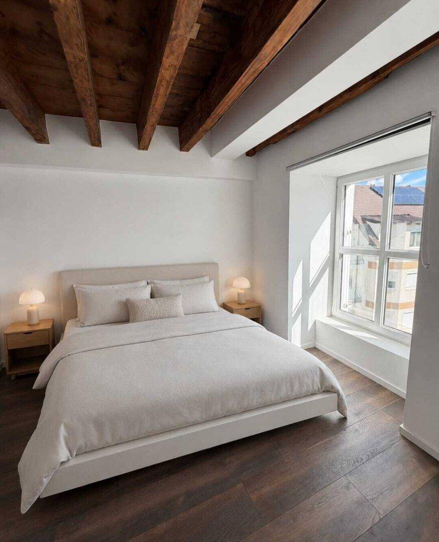 Helles Schlafzimmer mit Holzbalkendecke, großem Bett und Fenster mit Stadtblick.