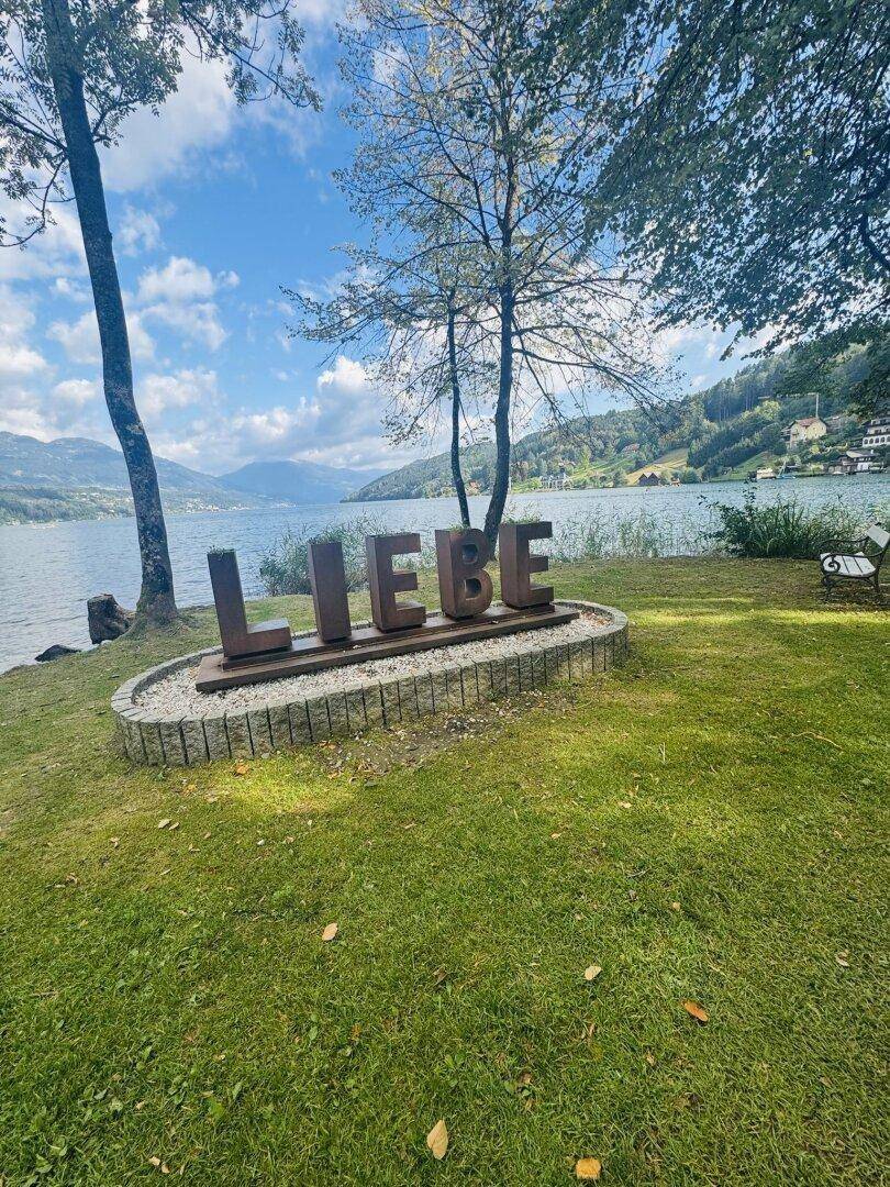 Große 'LIEBE'-Skulptur am Seeufer, umgeben von grüner Wiese und Bäumen, mit Seeblick im Hintergrund.