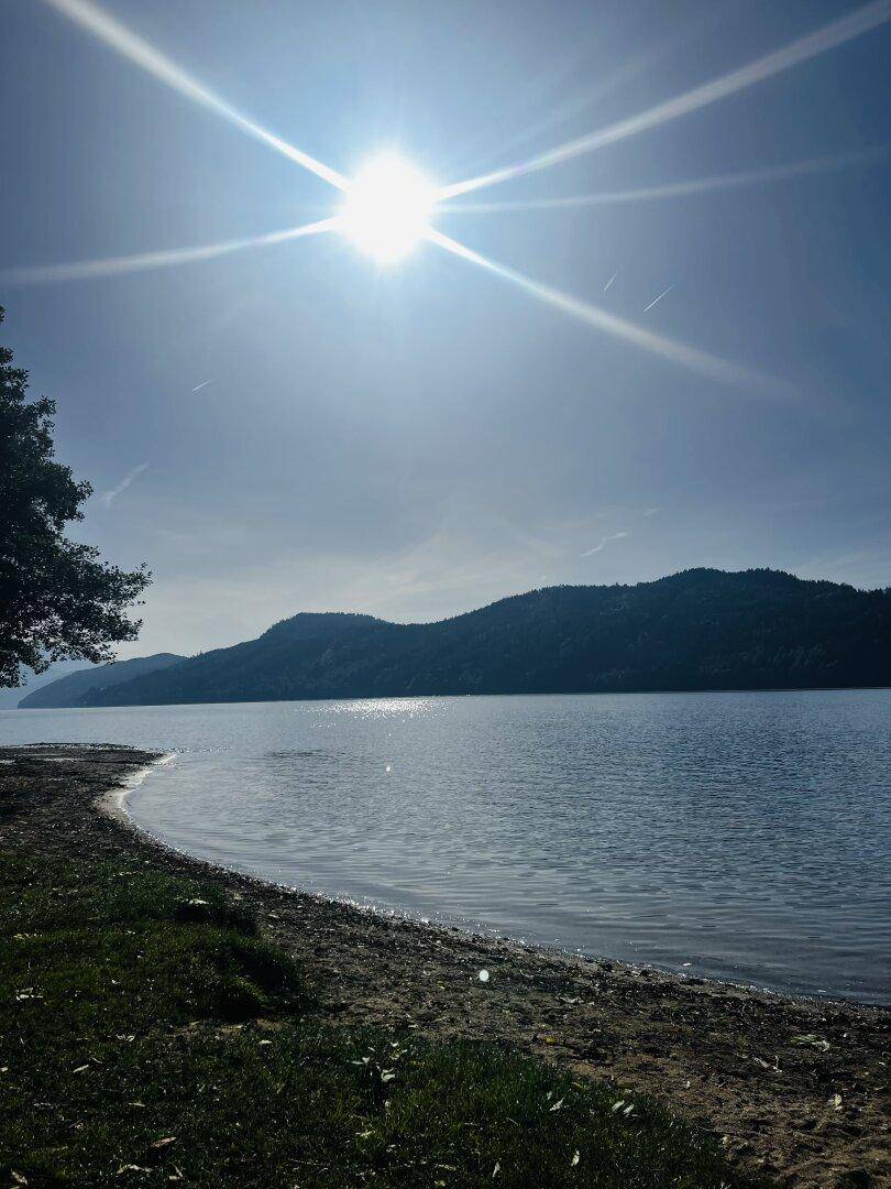 Sonniger Seeblick mit glitzerndem Wasser, umrahmt von bewaldeten Bergen und einem sandigen Uferbereich.