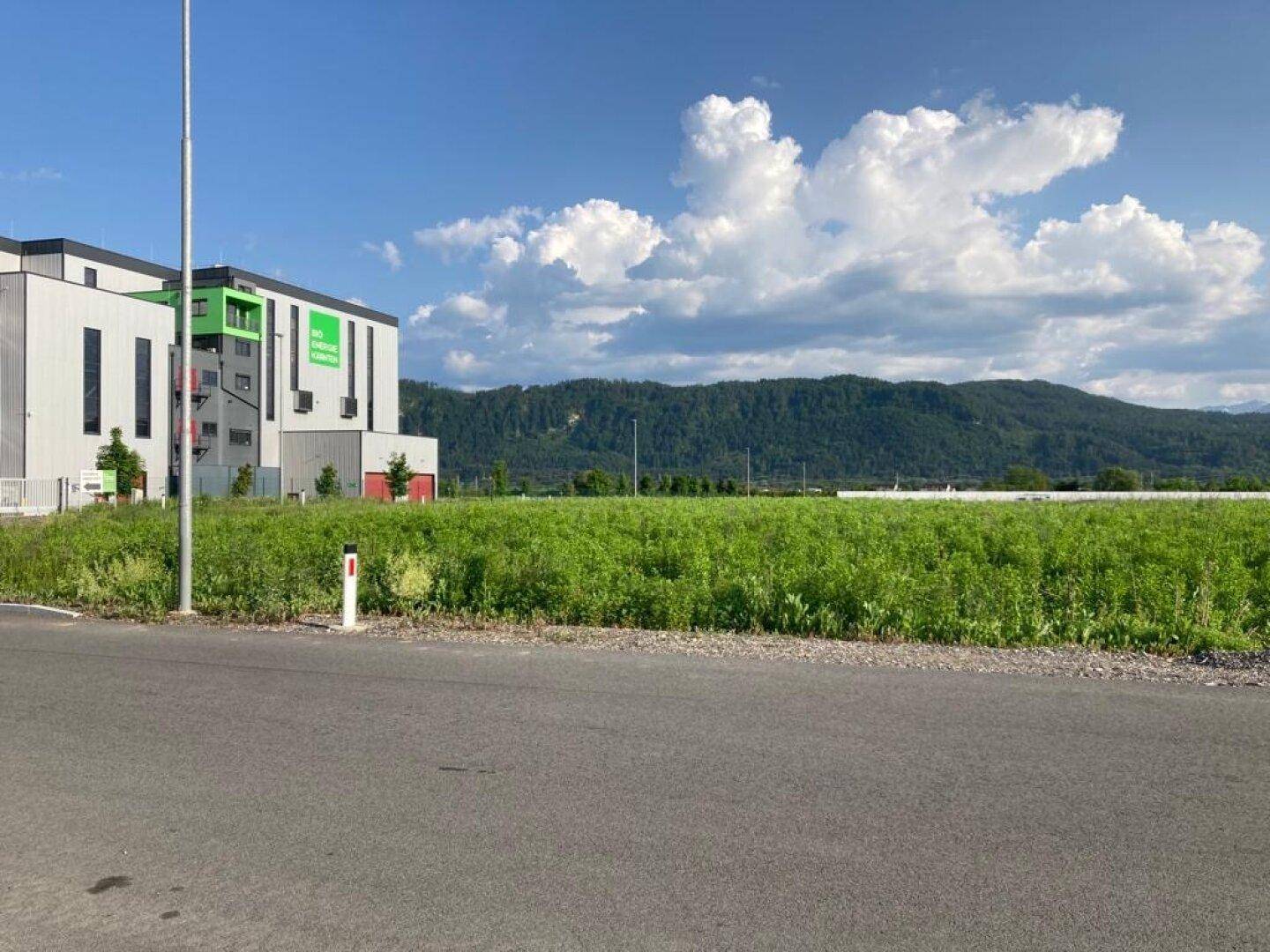 Modernes Gebäude mit grünen Akzenten vor einem weiten grünen Feld und einer Bergkulisse unter blauem Himmel.