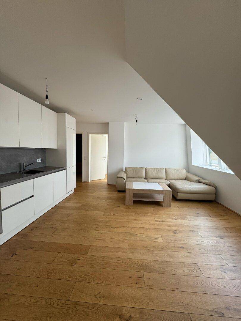 Heller offener Wohnbereich mit moderner Einbauküche, Sofa und Holzboden unter Dachschrägen.