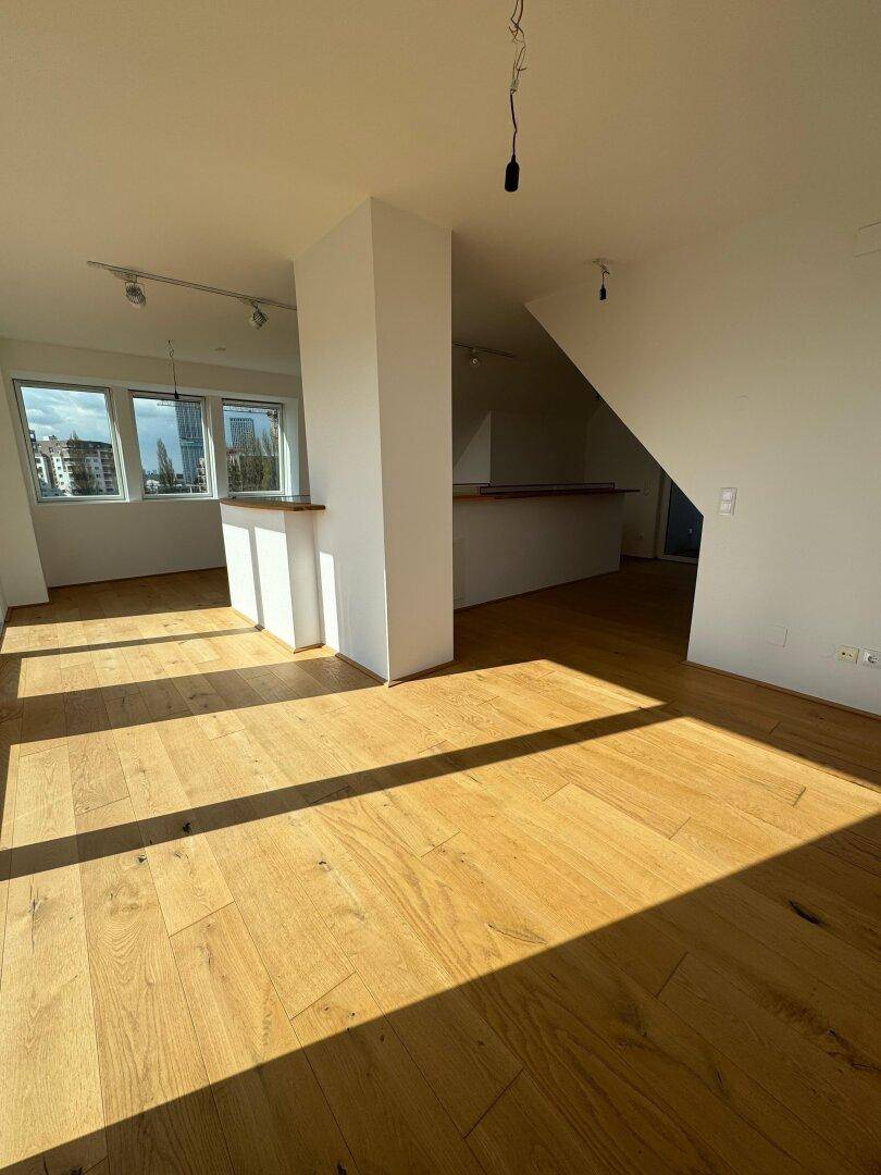 Geräumiger, lichtdurchfluteter Wohnbereich mit Holzboden und großen Fenstern, die viel Sonne hereinlassen.