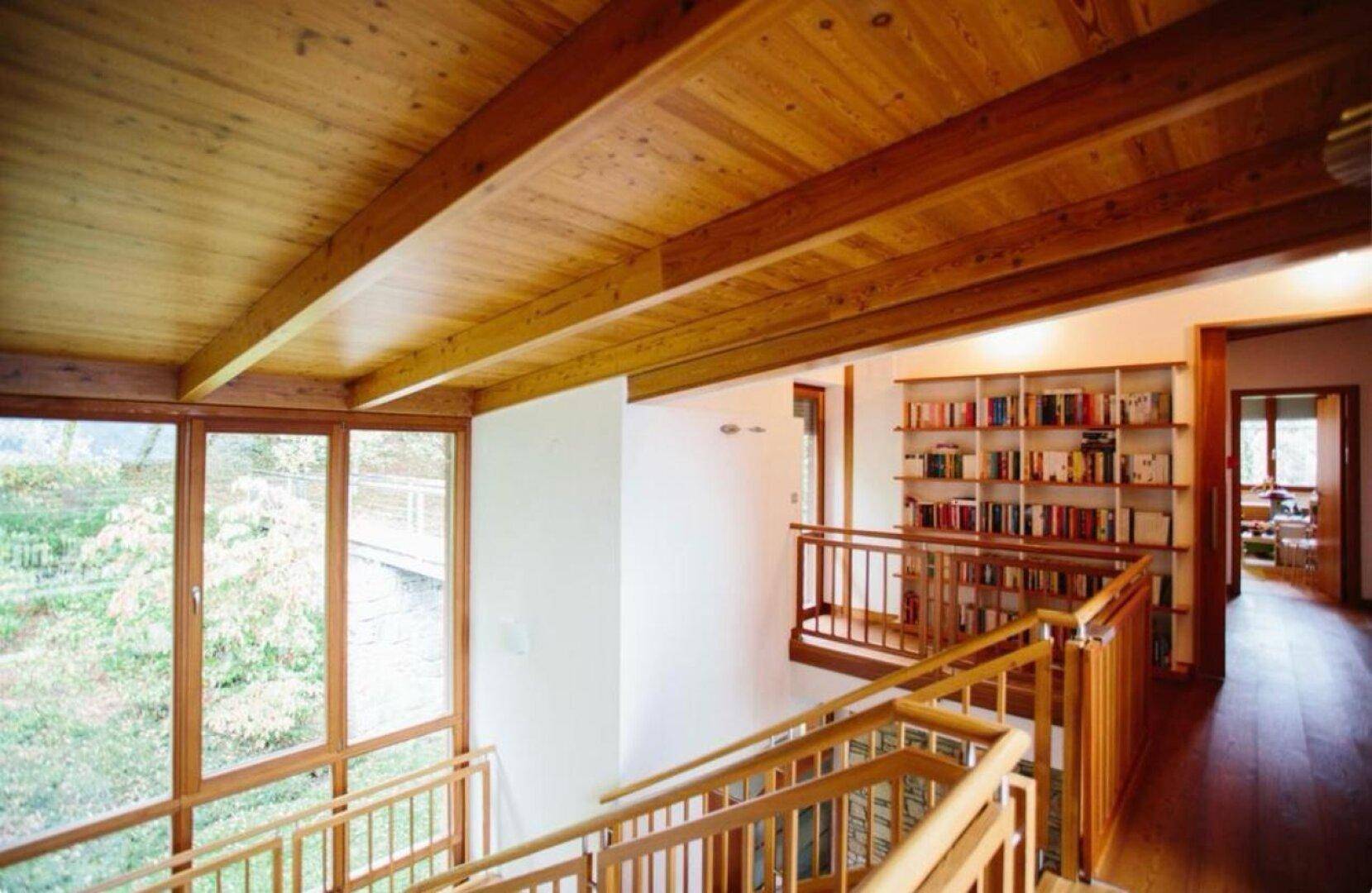 Heller Flur mit Holztreppe, hohen Decken und eingebautem Bücherregal, umgeben von großen Fenstern.