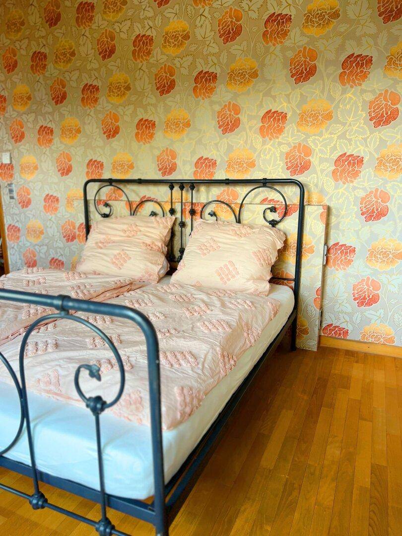 Schlafzimmer mit auffälliger floraler Tapete, schmiedeeisernem Bett und hellem Holzboden.