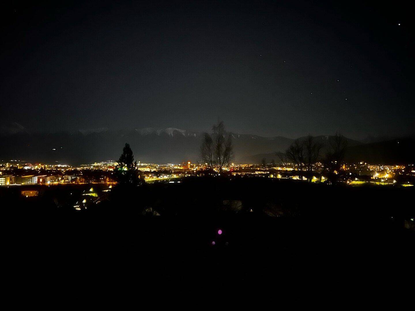 Panoramablick auf eine beleuchtete Stadt bei Nacht mit schneebedeckten Bergen im Hintergrund.