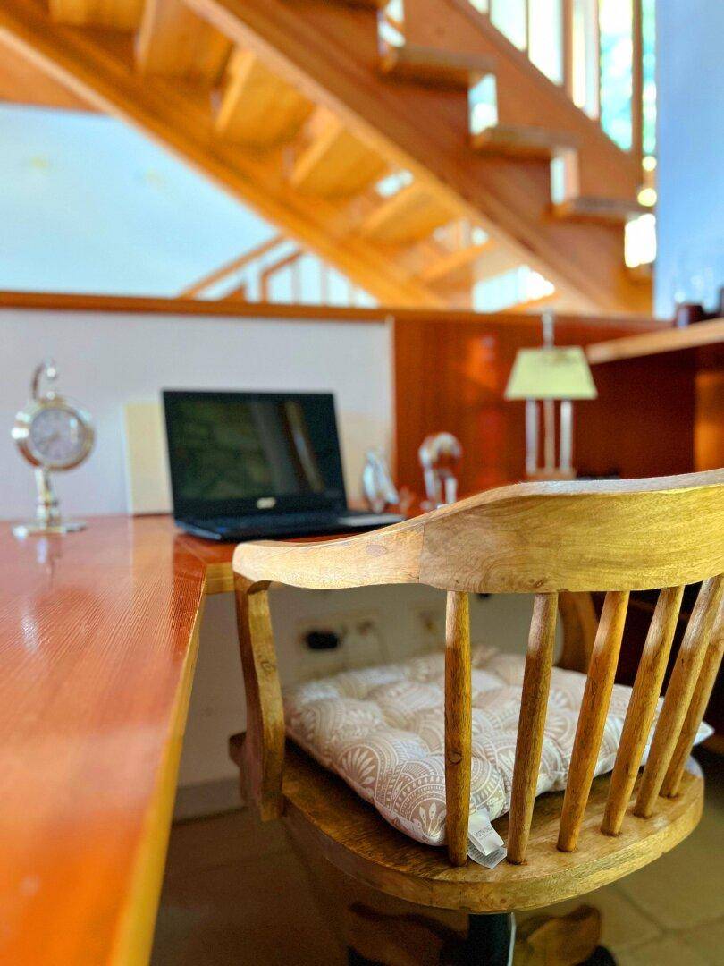 Heller Arbeitsplatz mit Holztisch und Laptop, unter einer Holztreppe gelegen, mit viel Tageslicht.