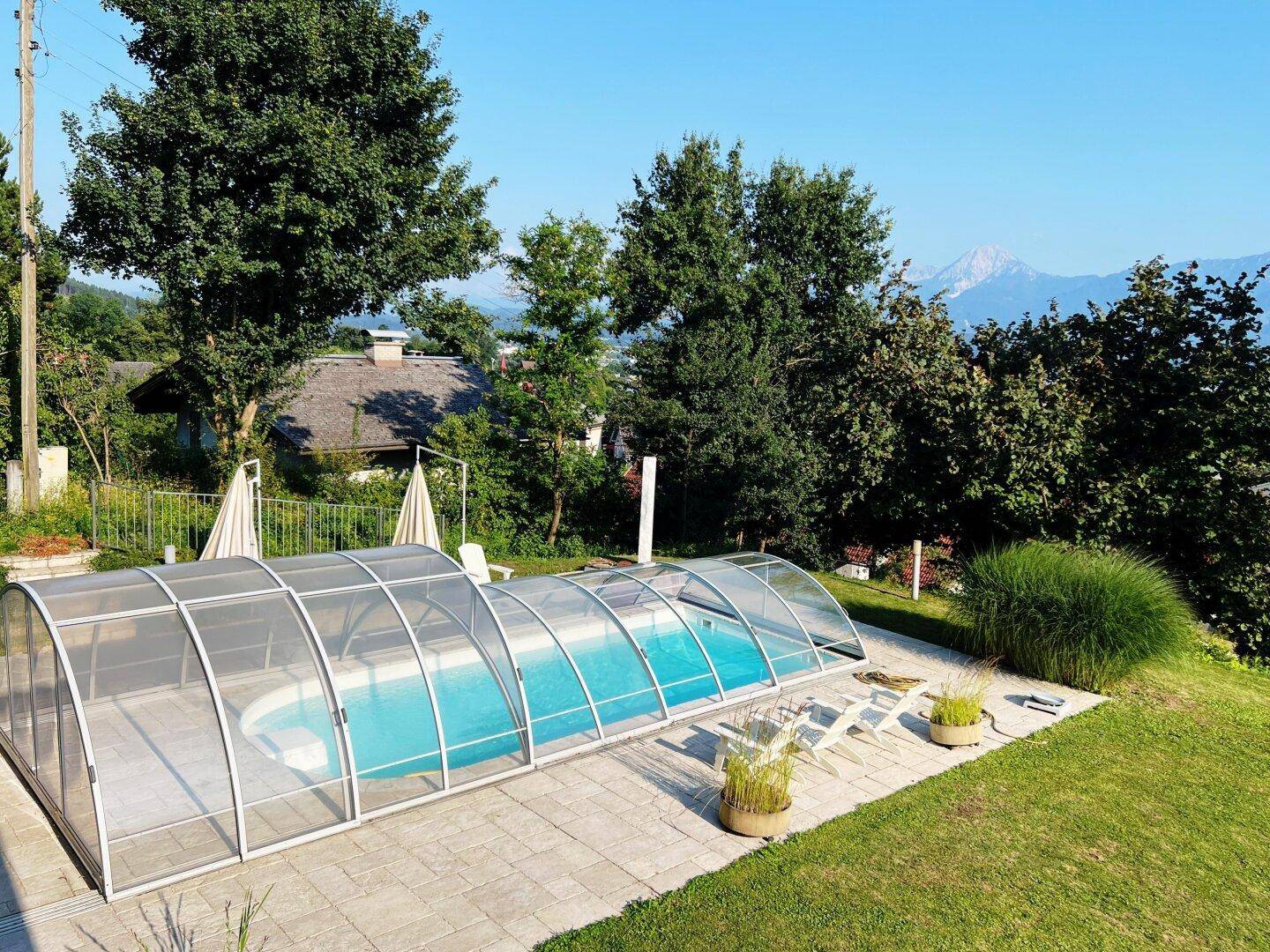 Gepflegter Garten mit überdachtem Pool und Liegeflächen, umgeben von Bäumen und mit Bergblick.
