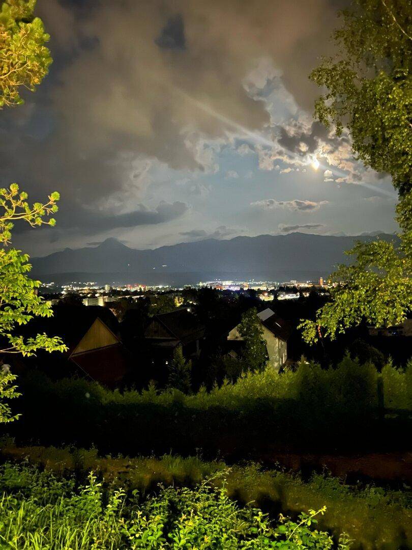 Nächtlicher Panoramablick auf eine Stadt mit Bergen im Hintergrund unter einem bewölkten Himmel mit Mond.