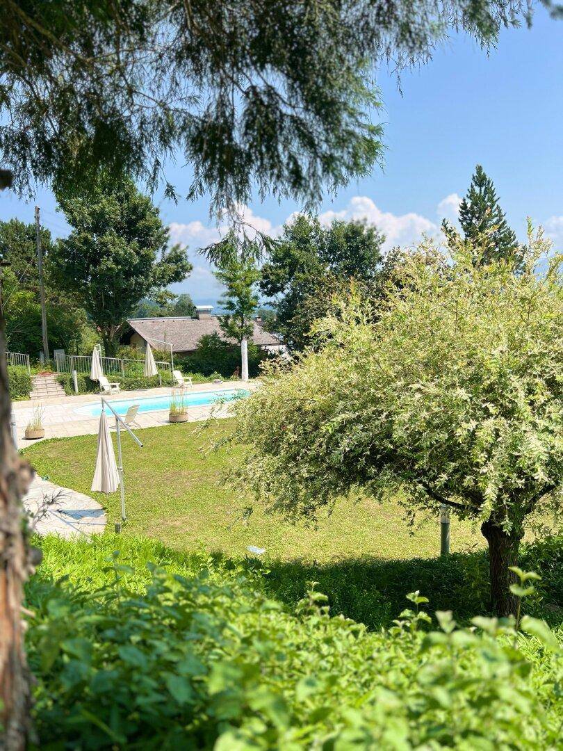 Weitläufiger Garten mit Rasenflächen, Bäumen und einem Poolbereich im Hintergrund unter blauem Himmel.