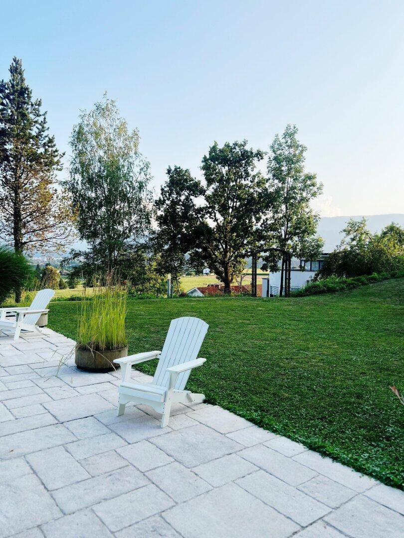 Sonniger Gartenbereich mit gepflasterter Terrasse, Rasenfläche und Adirondack-Stühlen zum Entspannen.