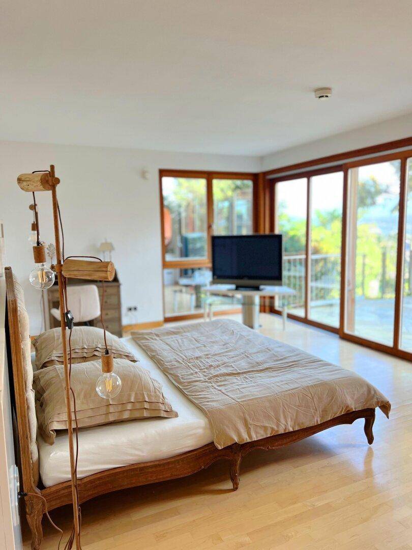 Helles Schlafzimmer mit Holzbett, großen Fenstern und Zugang zu einem Balkon oder einer Terrasse.