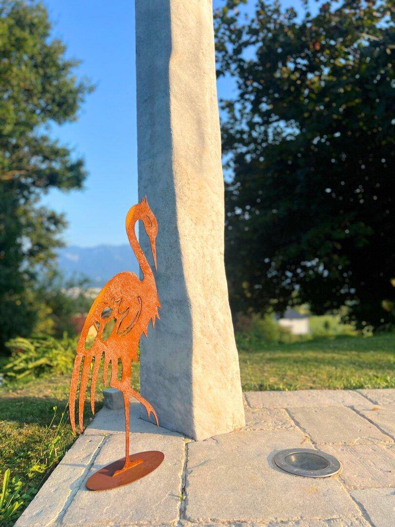 Dekorative Metallskulptur eines Kranichs in einem sonnigen Gartenbereich mit gepflastertem Weg.