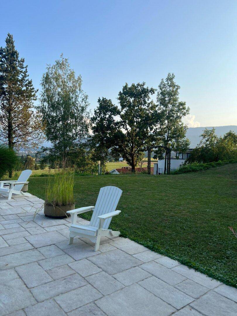 Gepflasterte Terrasse mit zwei weißen Adirondack-Stühlen, umgeben von Rasen und Bäumen.