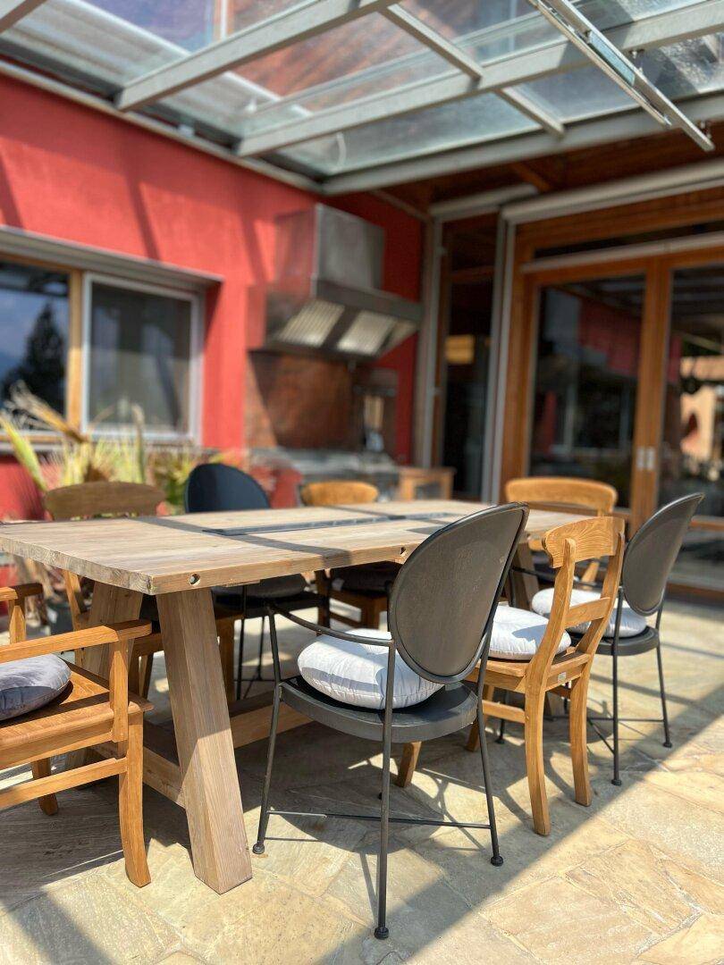 Großer Essbereich im Freien auf einer Terrasse mit Glasdach und roter Fassade.