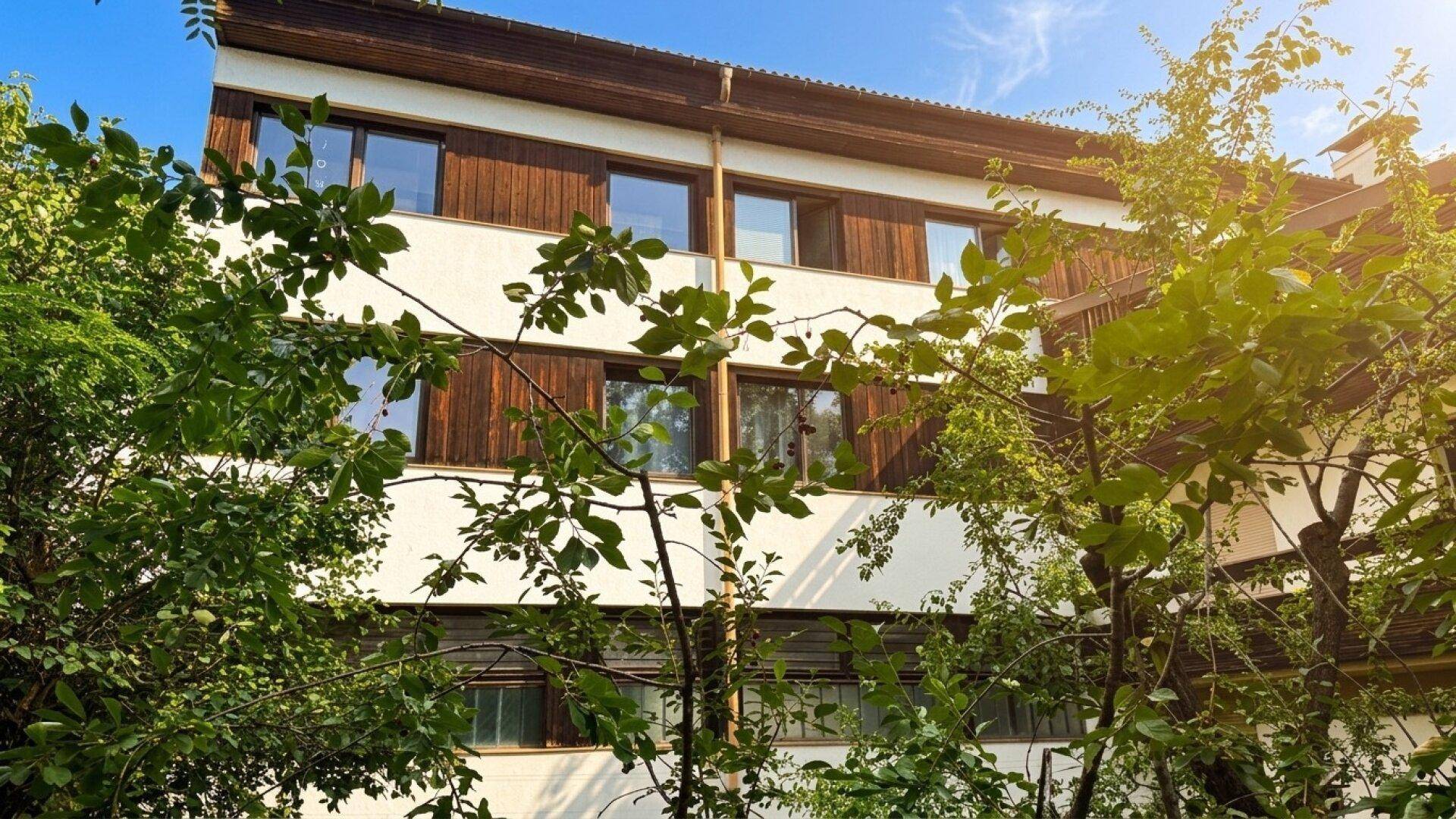 Gebäudefassade mit Holzverkleidung und weißen Abschnitten, teilweise von Bäumen verdeckt.