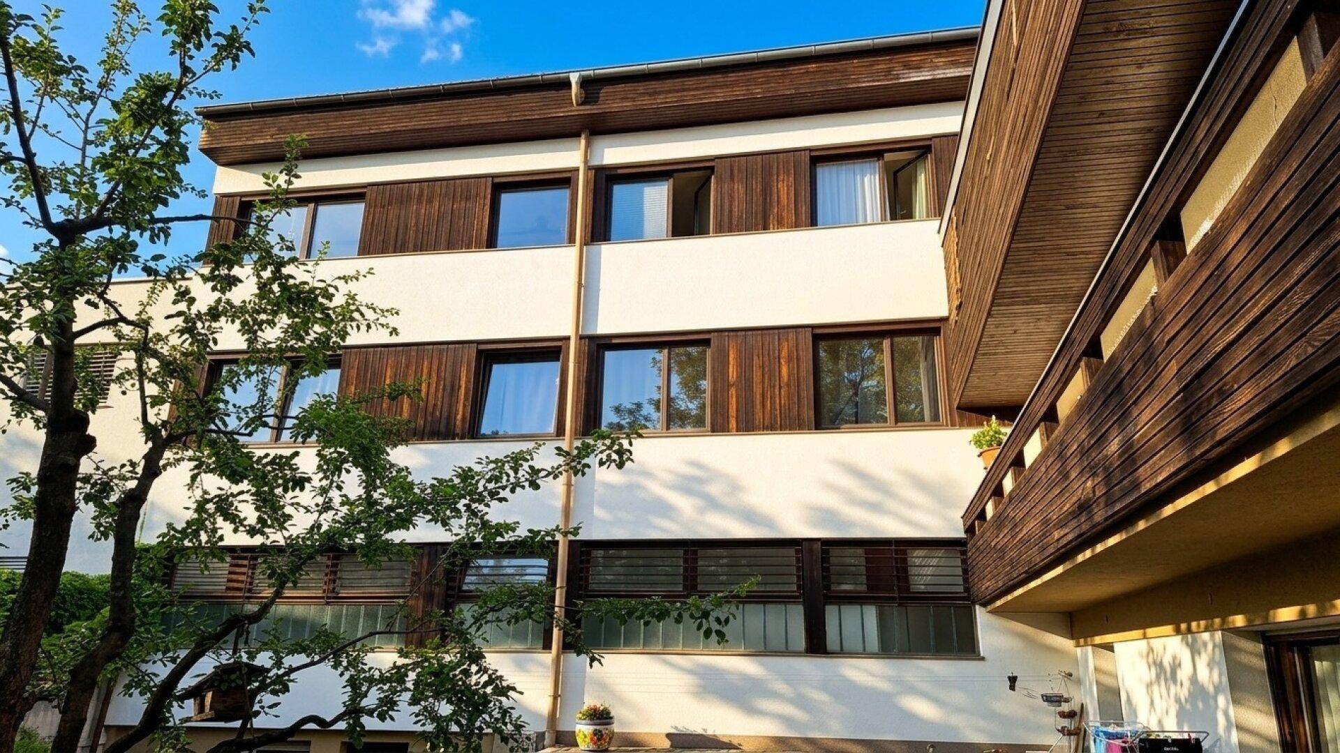 Gebäudefassade mit Holz- und Putzelementen, umgeben von grünen Bäumen unter blauem Himmel.