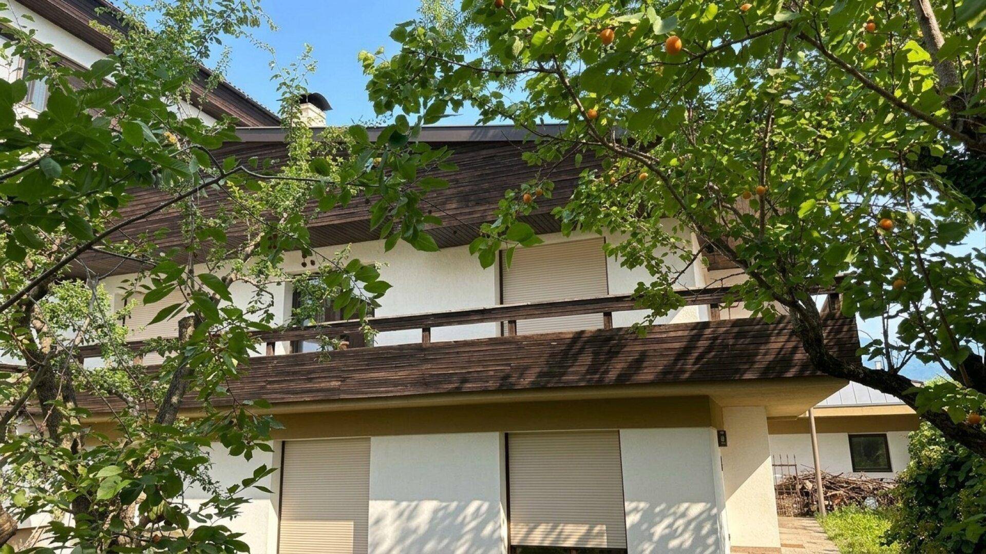 Gebäudefassade mit einem Balkon und Rollläden, umgeben von üppigem Grün und Bäumen.