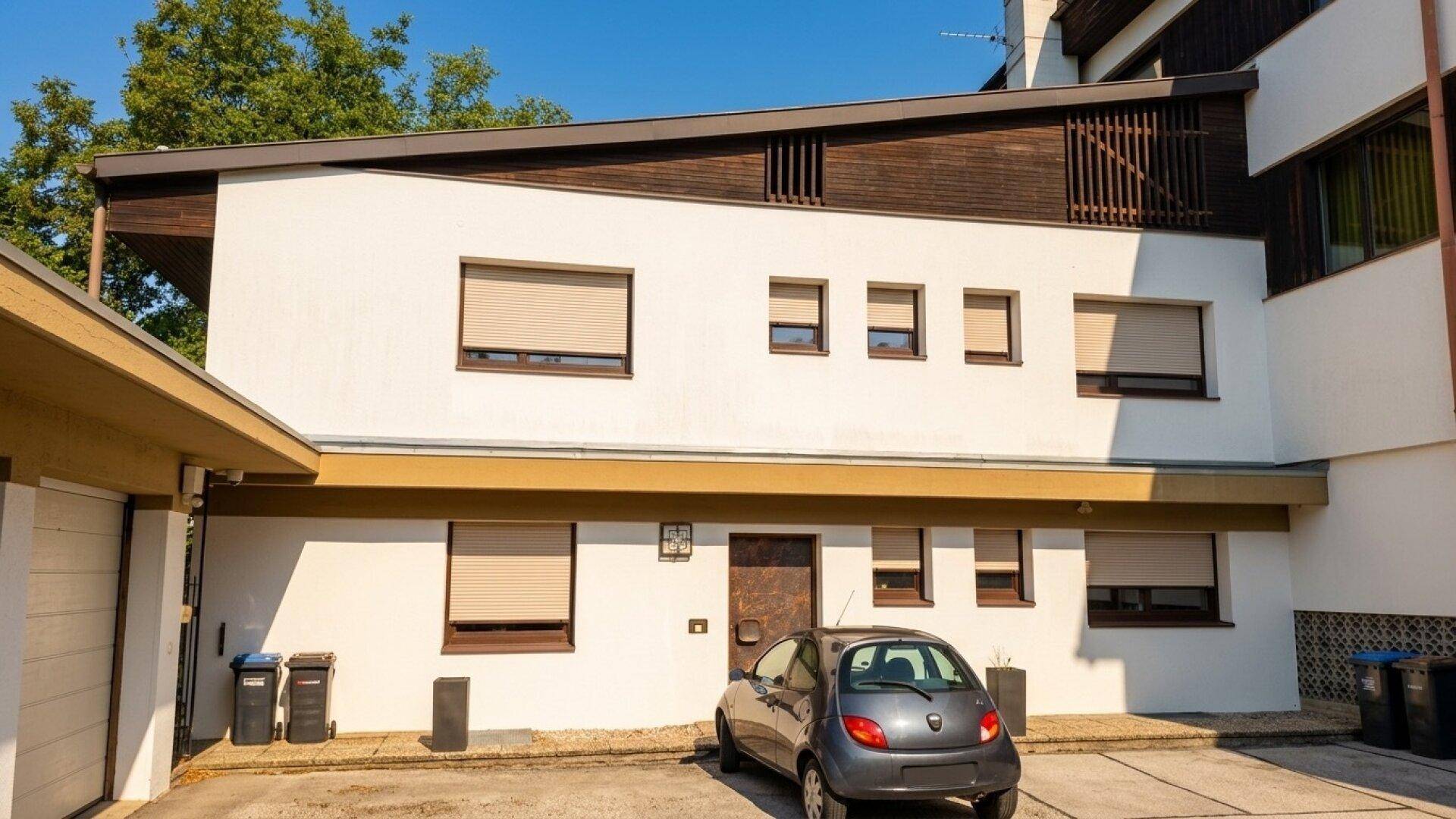 Gebäudefassade mit Rollläden an den Fenstern und einem gepflasterten Parkplatz davor.