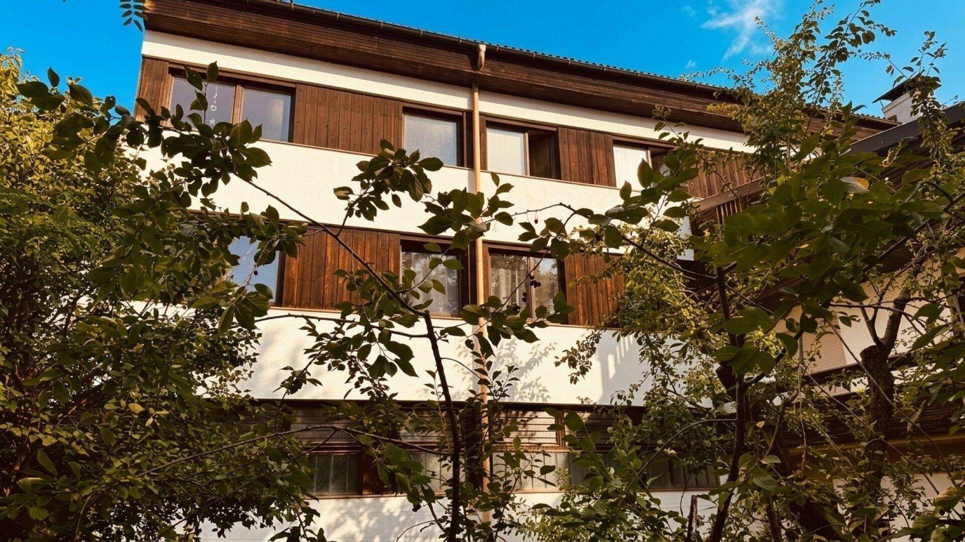 Fassade eines Gebäudes mit Holzverkleidung und weißen Abschnitten, teilweise von Bäumen verdeckt.