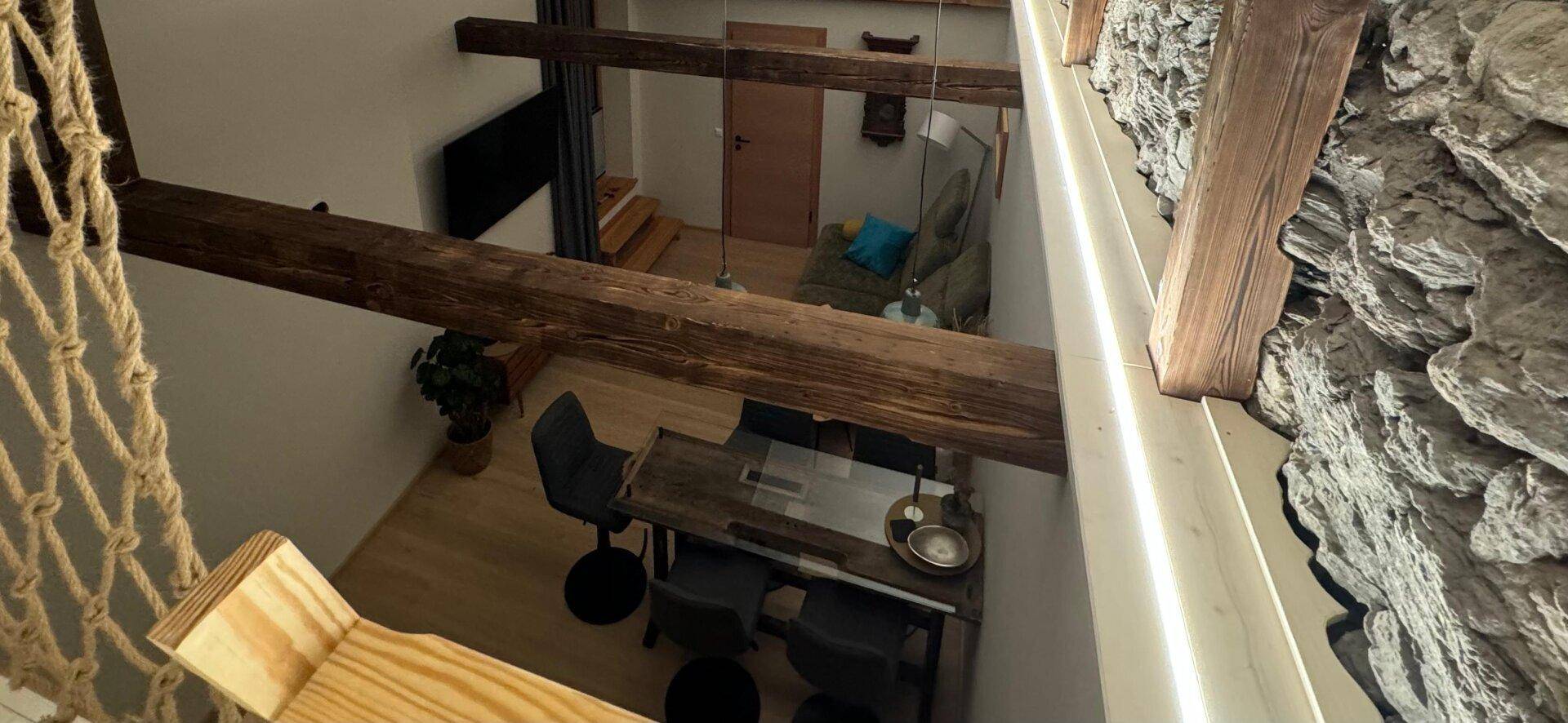 Blick von oben auf den offenen Wohn-Essbereich mit Holzbalken und einer rustikalen Steinwand, die den Loft-Charakter unterstreicht.