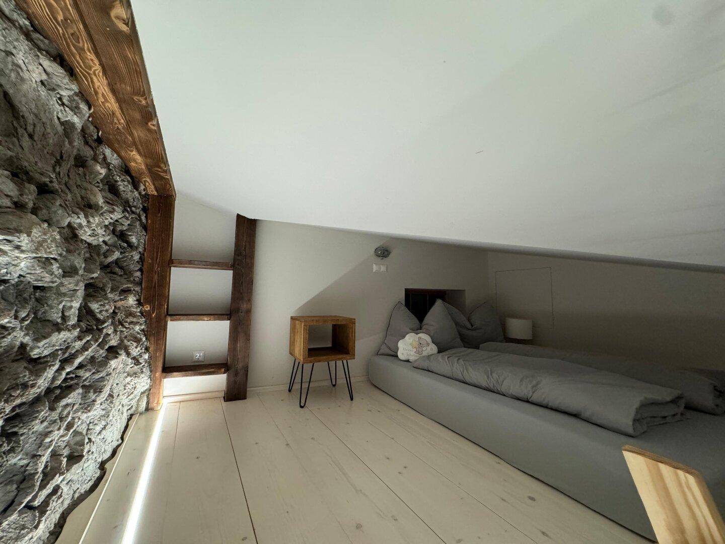 Gemütlicher Schlafbereich im Dachgeschoss mit niedriger Decke, Holzboden und einer rustikalen Steinwand.