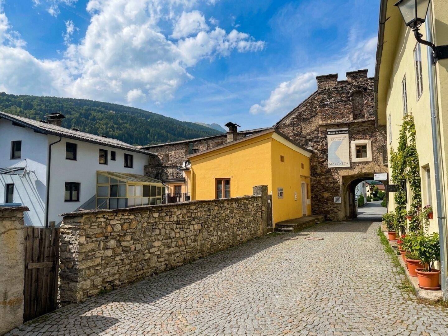 Historische Gasse mit Kopfsteinpflaster, alten Steinmauern und Blick auf die umliegende Berglandschaft unter blauem Himmel.