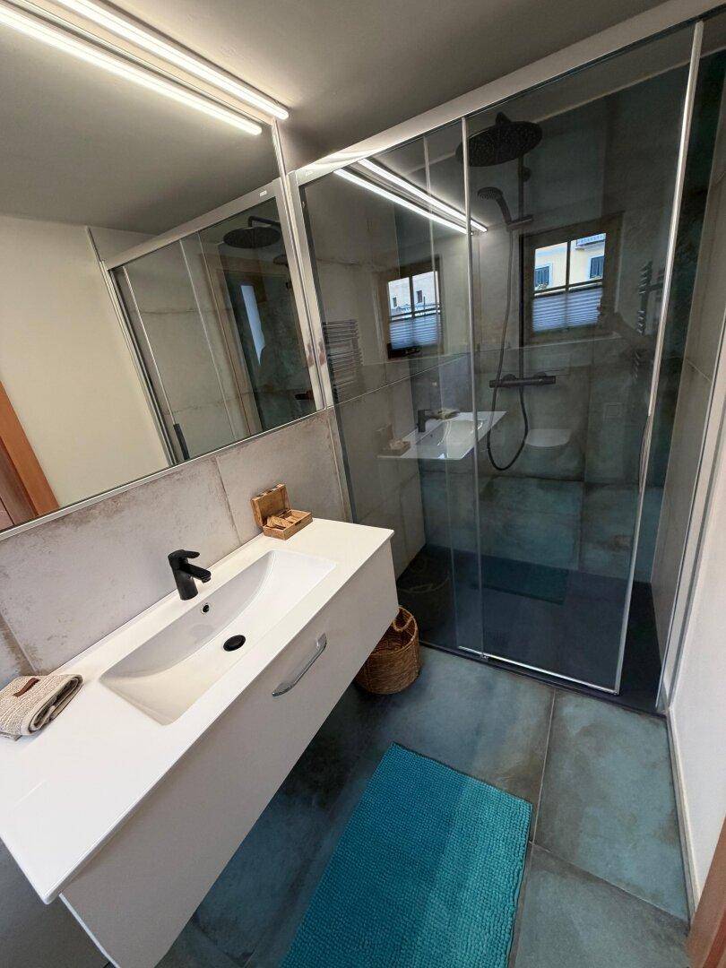 Modernes Badezimmer mit begehbarer Dusche, Einzelwaschbecken und großformatigen Fliesen in Grau- und Blautönen.