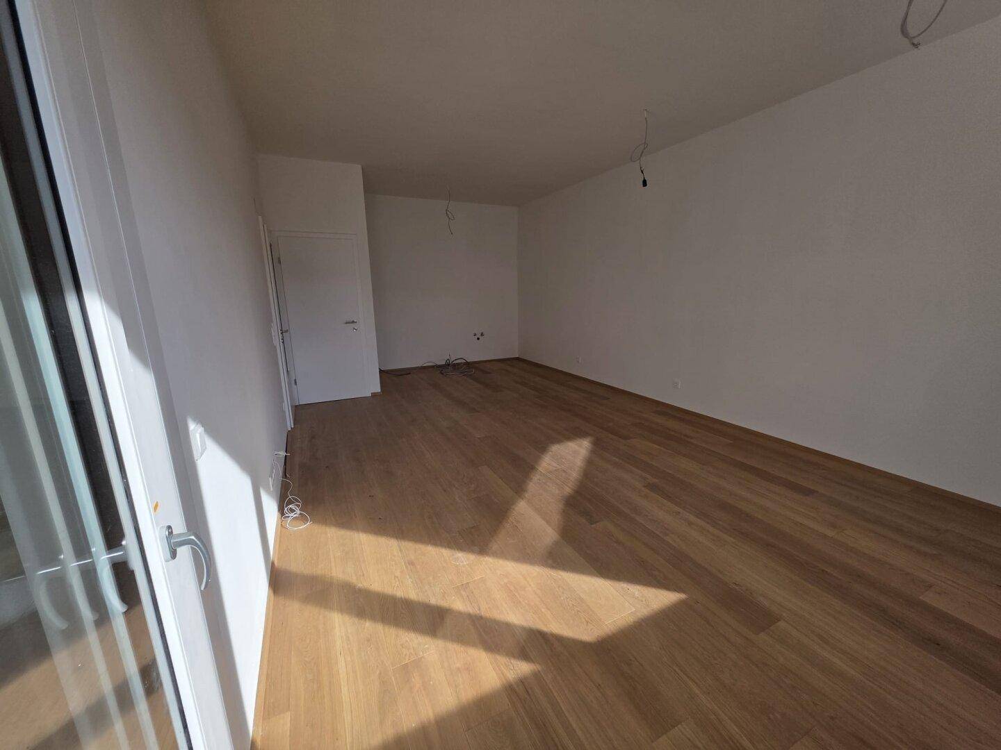 Leerer Raum mit hellem Holzboden, weißen Wänden und viel Tageslicht.