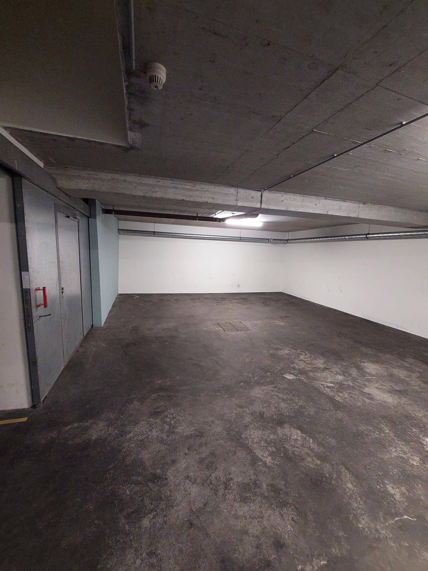 Leerer Tiefgaragenstellplatz mit Betonboden, unverputzter Decke und Metalltüren.