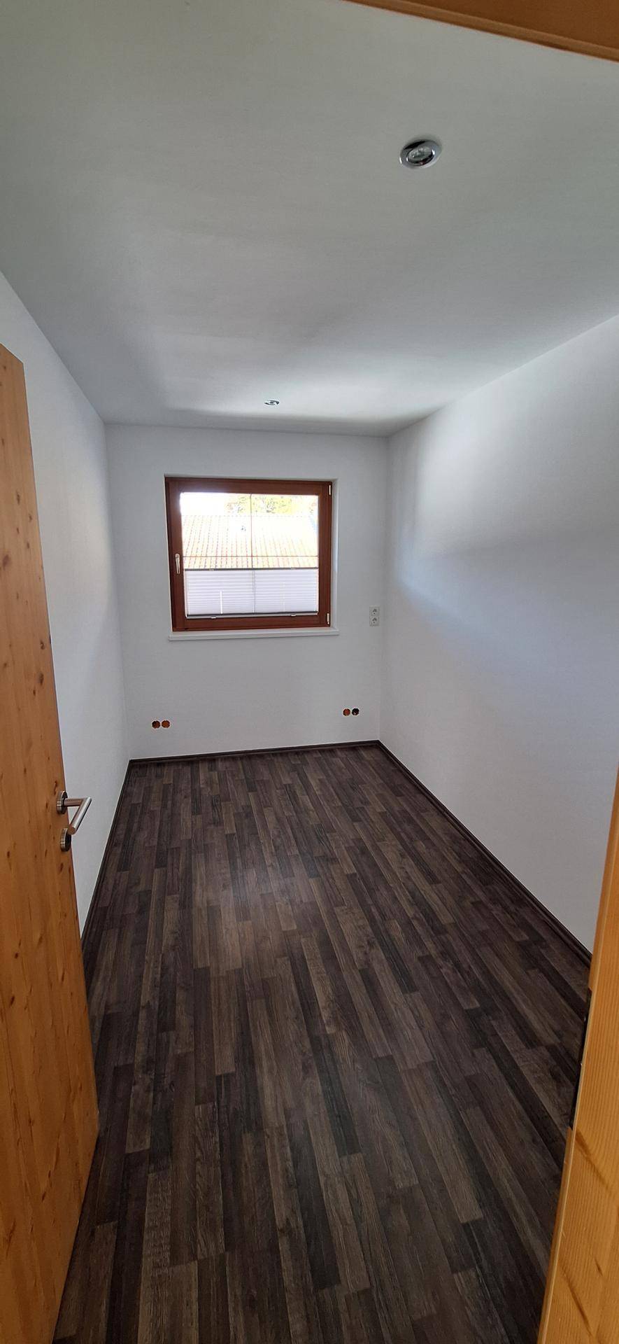 Helles Zimmer mit dunklem Laminatboden und einem Fenster, ideal als Kinder- oder Gästezimmer.