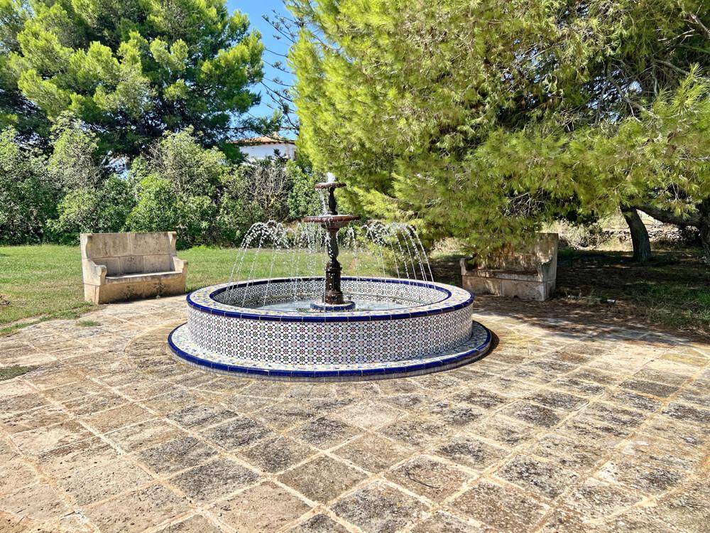 Mediterraner Brunnen mit farbigen Fliesen und sprudelndem Wasser, umgeben von Bäumen und Steinplatten.