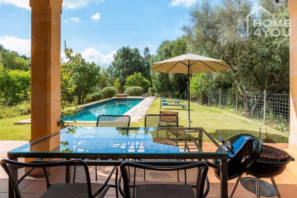 Überdachte Terrasse mit Esstisch, Grill und Blick auf den Pool und die grüne Umgebung.